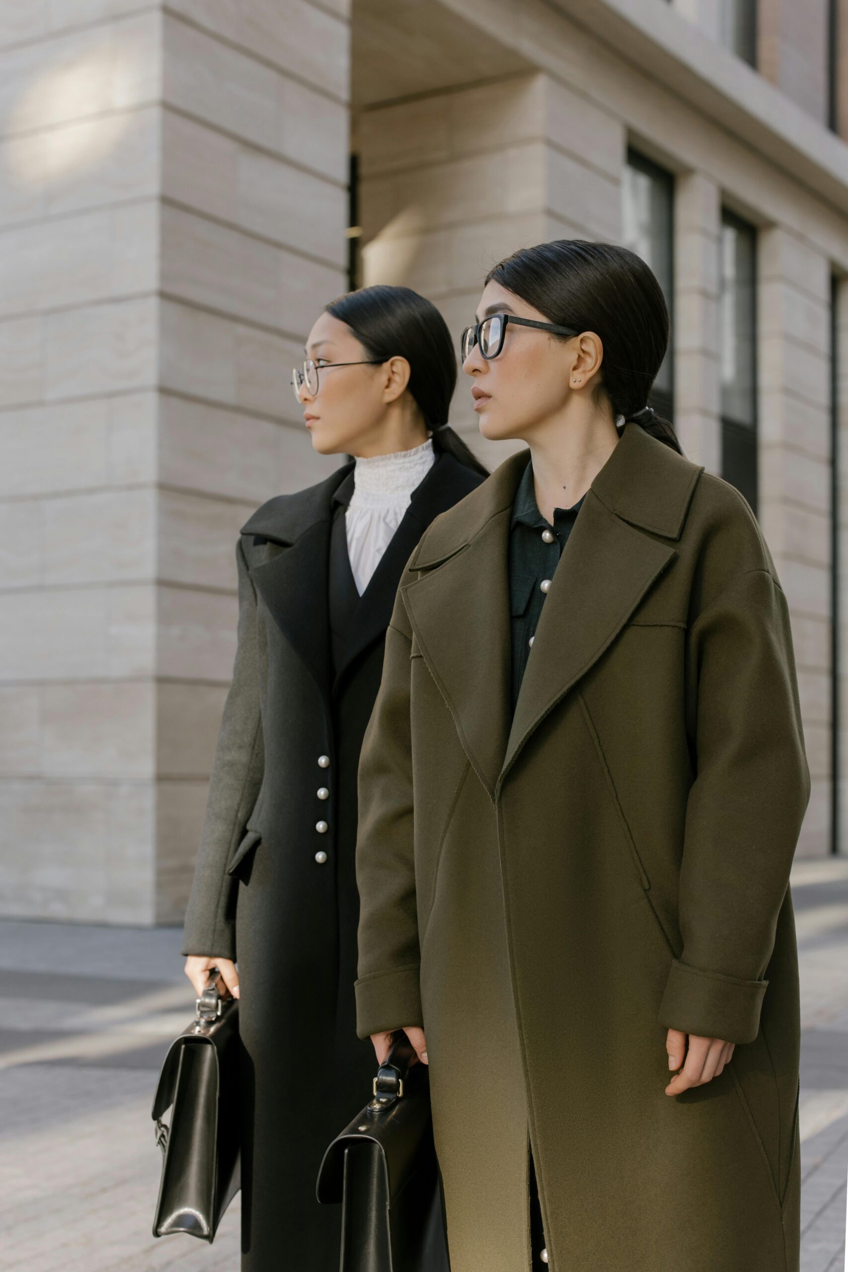 Two stylish women in elegant coats standing outdoors with briefcases, showcasing urban fashion.