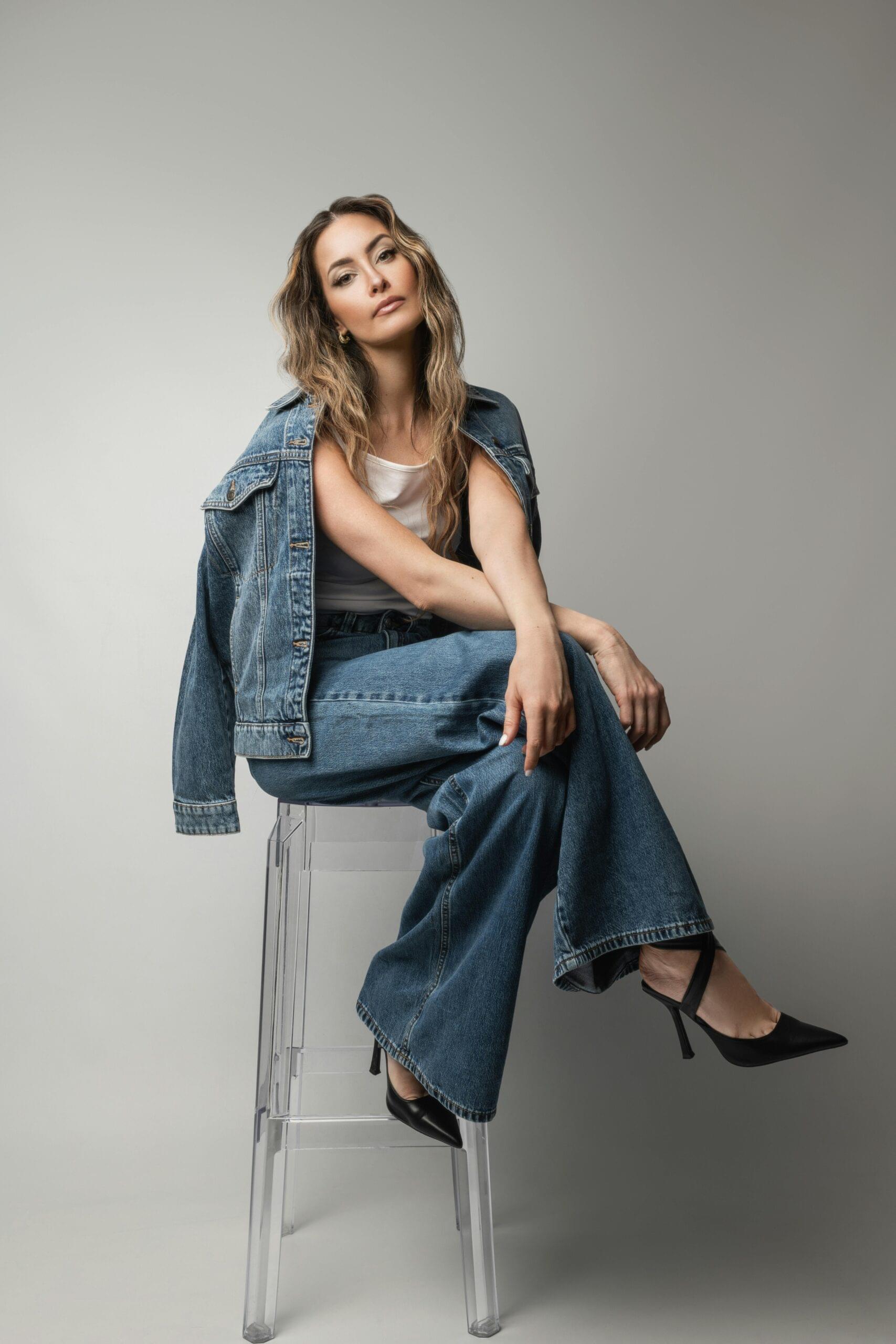 Fashionable woman wearing denim jacket and jeans sitting on a stool. Perfect for lifestyle and fashion concepts.